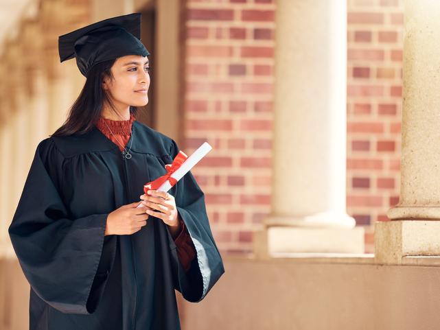 A woman in a mortarboard, holding a scroll, looks off into the distance