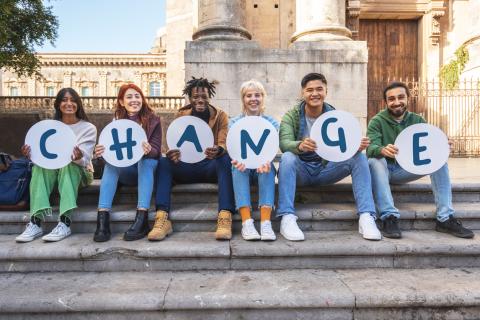 Students sitting on the steps of a campus building each holding a paper with a letter of the word "change" on it