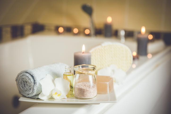 A tray with a jar of bath salts, lilies and towels sits on the edge of a candelit bathtub