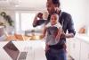 A man with a baby in a sling checks his laptop while drinking coffee